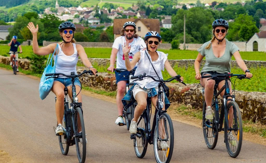 4 cyclistes faisant des signes de la main au photographe. Ils roulent sur une route travers les vignes