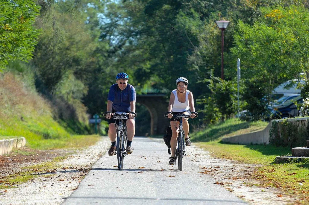 Deux cyclistes roulant sur une piste cyclable. Il y'a au fond un grand pont en pierre