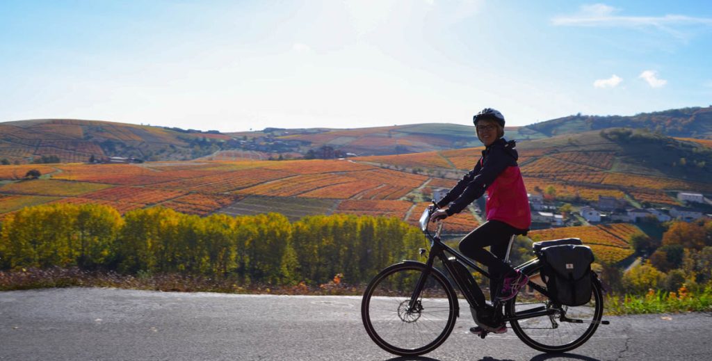 Cyclist smiling at the camera. Behind her, endless vineyards, in orange and yellow tones.